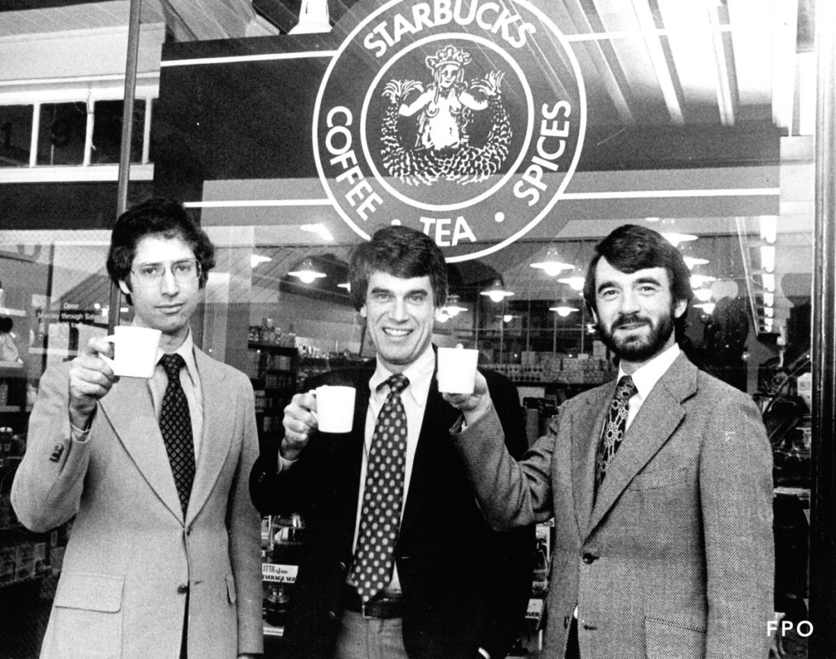 Foto em preto e branco de 1979, com Zev Siegl, Jerry Baldwin and Gordon Bowker de terno, em frente à Starbucks e com uma xícara de café em mãos.