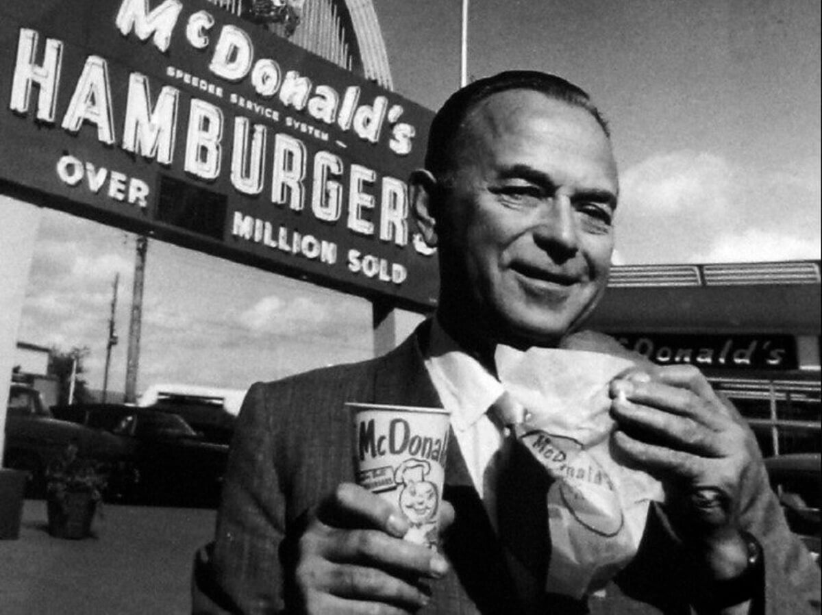 Ray Kroc, co fundador do mcdonalds em frente a um restaurante da rede, comendo um hamburguer