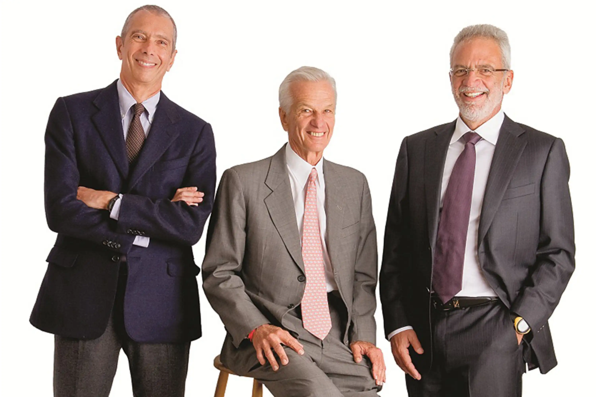 Carlos Alberto Sicupira em pé com os braços cruzados, Jorge Paulo Lemann sentado e Marcel Herrmann Telles em pé com a mão no bolso. Todos estão vestindo ternos.